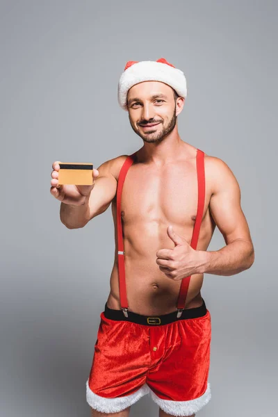 Musculoso hombre en sombrero de Navidad haciendo pulgar hacia arriba y mostrando la tarjeta de crédito aislado sobre fondo gris - foto de stock
