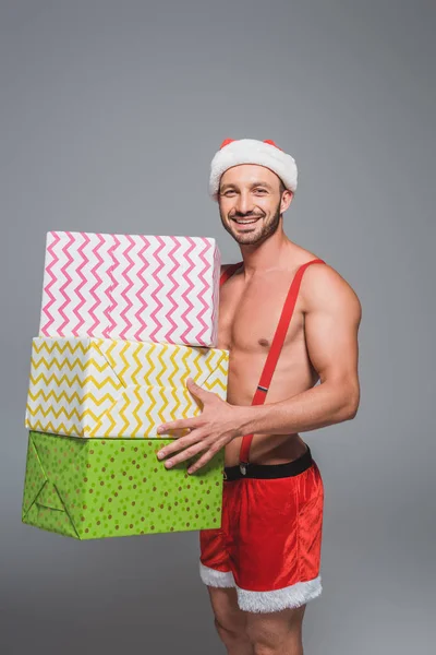 Счастливый мускулистый человек в рождественской шляпе с кучей подарочных коробок, изолированных на сером фоне — стоковое фото