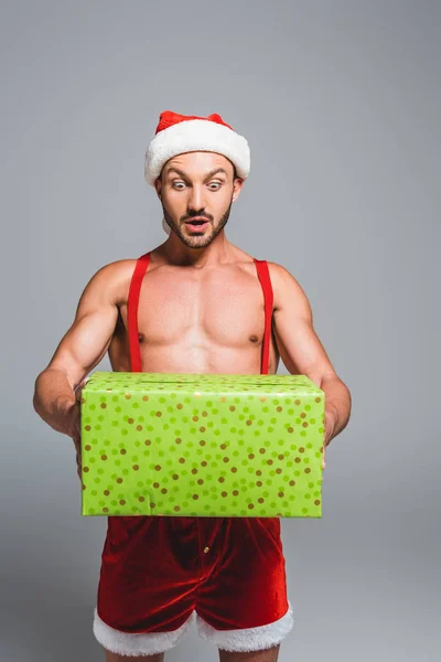 Chocado muscular homem no Natal chapéu segurando caixa de presente isolado no fundo cinza — Fotografia de Stock