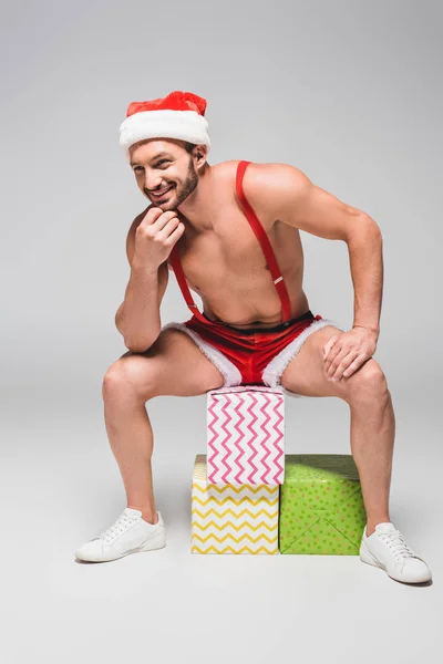 Homem muscular sorridente em chapéu de Natal e shorts sentado em caixas de presente em fundo cinza — Fotografia de Stock