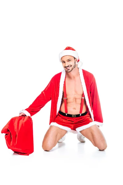 Sonriente musculoso hombre en santa claus traje sentado con navidad saco aislado en blanco fondo - foto de stock