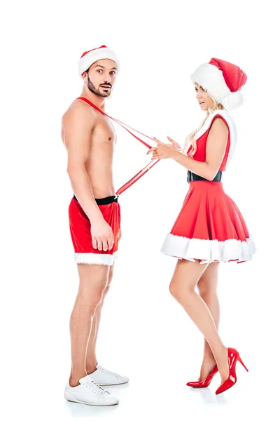 Side view of young woman in santa hat and dress pulling susenders of her shocked boyfriend isolated on white background — стоковое фото