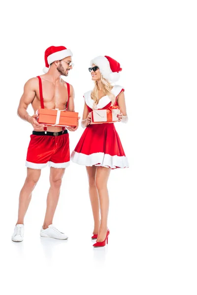 Pareja joven en sombreros de Navidad y gafas de sol sosteniendo cajas de regalo y mirándose aislados sobre fondo blanco - foto de stock