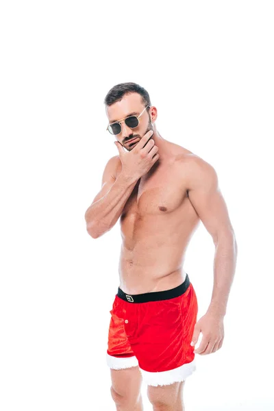 Confiante shirtless muscular homem em óculos de sol e calções de Papai Noel de pé isolado no fundo branco — Fotografia de Stock