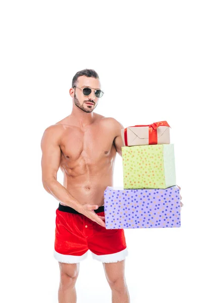 Uomo muscoloso senza maglietta in occhiali da sole e pantaloncini di Babbo Natale tenendo mucchio di regali isolati su sfondo bianco — Foto stock