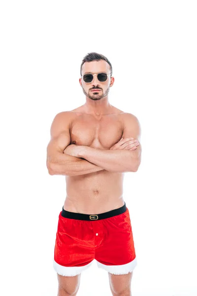Hombre musculoso serio en pantalones cortos de Navidad y gafas de sol de pie con brazos cruzados aislados sobre fondo blanco - foto de stock