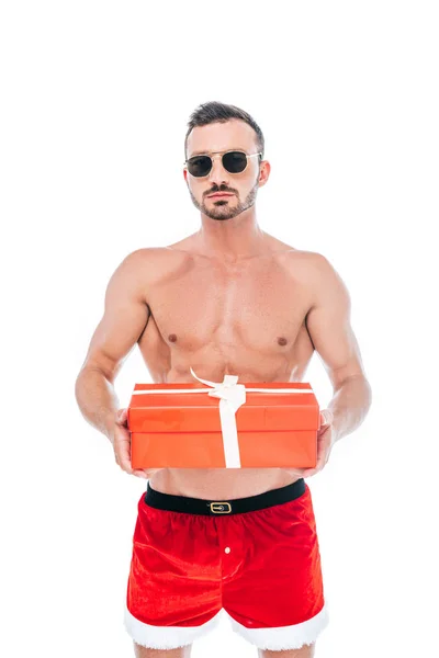 Grave shirtless muscular homem em santa shorts e óculos de sol segurando caixa de presente isolado no fundo branco — Fotografia de Stock