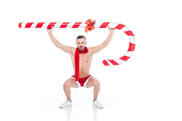 Hemdloser muskulöser Mann in Weihnachtsmann-Shorts und rotem Schal, der mit großen gestreiften Weihnachtsstäben auf weißem Hintergrund übt — Stockfoto