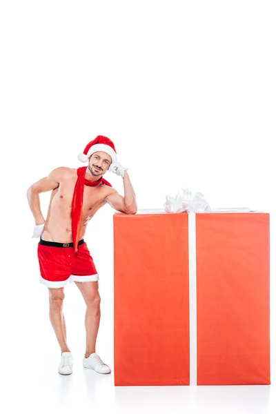 Junger, muskulöser Mann mit Weihnachtsmütze, der in der Nähe einer großen Geschenkbox auf weißem Hintergrund steht — Stockfoto