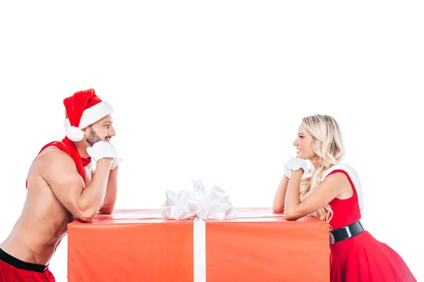Вид сбоку мечтательной пары в рождественской одежде, смотрящей друг на друга возле большой подарочной коробки, изолированной на белом фоне — стоковое фото