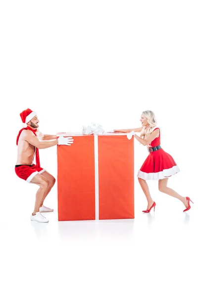 Vista lateral de feliz pareja en ropa de Navidad tirando de la caja de regalo grande en diferentes direcciones aisladas sobre fondo blanco - foto de stock