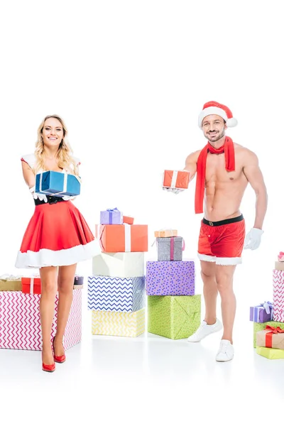 Attractive young woman in santa dress and muscular shirtless man in christmas hat giving presents isolated on white background — Stock Photo