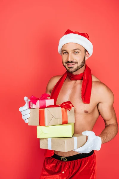 Glücklich muskulöse hemdlose Mann mit Weihnachtsmütze und rotem Schal hält Stapel von Geschenkboxen isoliert auf rotem Hintergrund — Stockfoto