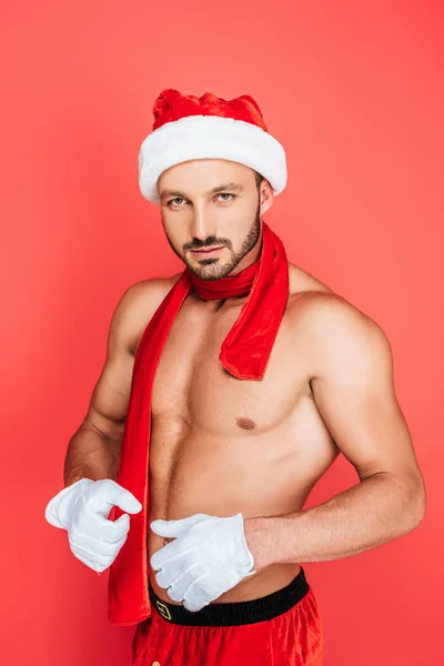 Confiado hombre musculoso sin camisa en sombrero de Navidad y guantes blancos mirando cámara aislada sobre fondo rojo - foto de stock