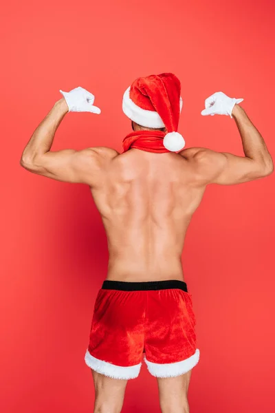 Vista trasera del hombre musculoso sin camisa en sombrero de Navidad y pantalones cortos que muestran los músculos aislados en el fondo rojo - foto de stock