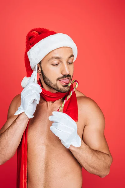 Muscular shirtless man in christmas hat and red scarf talking on smartphone and licking christmas stick isolated on red background — Stock Photo