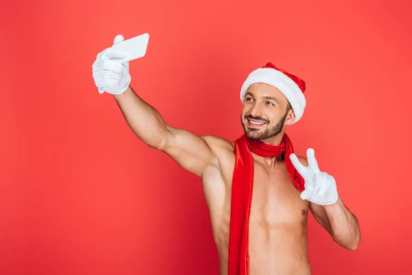 Smiling muscular shirtless man in christmas hat and red scarf taking selfie on smartphone and doing peace sign isolated on red background — Stock Photo