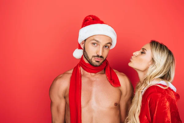 Vista lateral de mujer joven con los ojos cerrados tratando de besar a su novio en la bufanda sombrero de Navidad aislado sobre fondo rojo - foto de stock