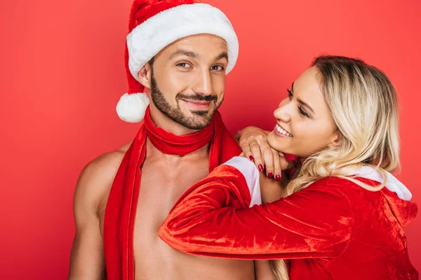 Портрет счастливого человека без рубашки в рождественской шляпе и красном шарфе и его улыбающейся подруги, стоящей рядом изолированной на красном фоне — стоковое фото
