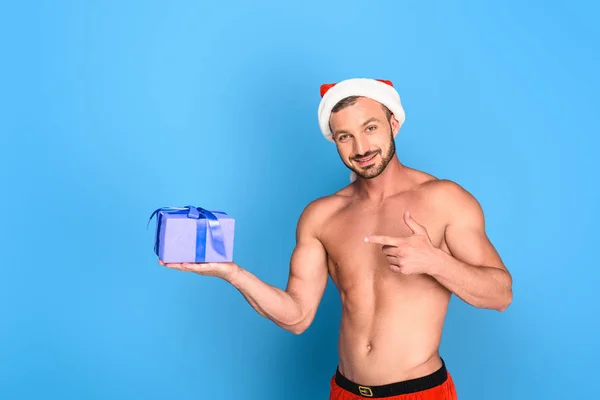 Handsome shirtless muscular man in christmas hat pointing at gift box isolated on blue background — Stock Photo