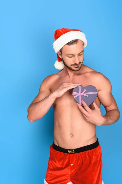 Handsome shirtless muscular man in christmas hat showing gift box isolated on blue background — Stock Photo