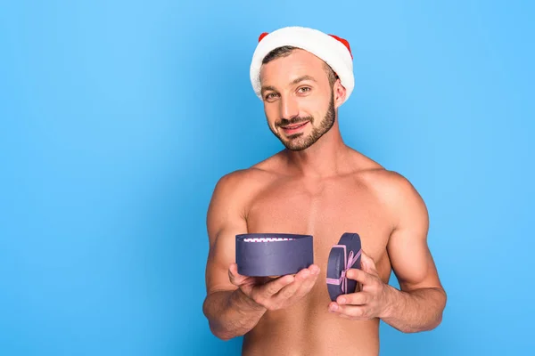 Portrait of shirtless muscular man in christmas hat holding gift box isolated on blue background — Stock Photo