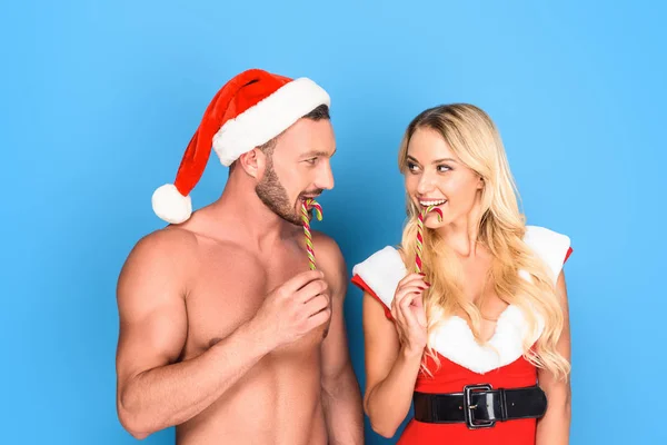 Happy boyfriend in christmas hat and girlfriend santa dress licking striped christmas sticks isolated on blue background — Stock Photo