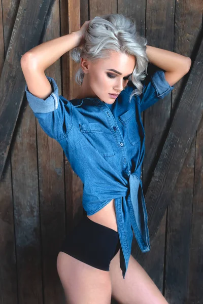 Belle jeune femme en denim chemise et culotte debout devant la porte en bois rustique et regardant la caméra — Photo de stock