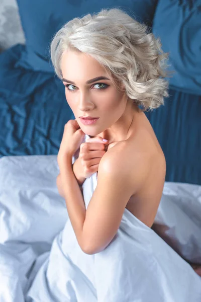 Hochwinkelaufnahme der attraktiven nackten jungen Frau, die ihren Körper mit einer Decke bedeckt, während sie im Bett sitzt und in die Kamera schaut — Stockfoto