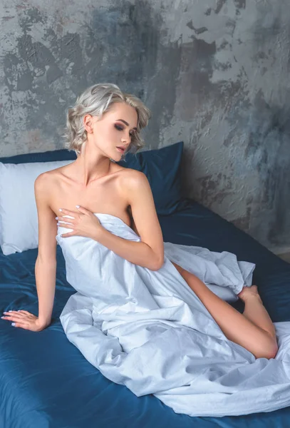 Seductive naked young woman sitting on bed and covering her body with blanket — Stock Photo