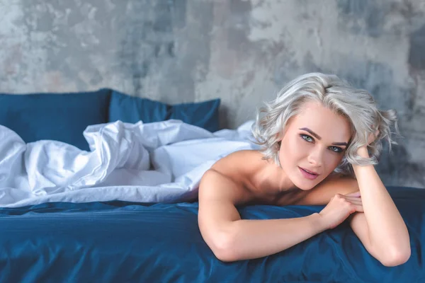 Atractiva mujer joven desnuda acostada en la cama debajo de la manta y mirando a la cámara - foto de stock