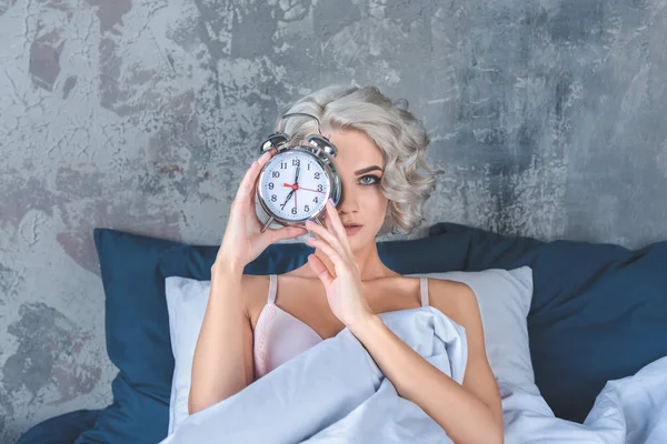 Привлекательная молодая женщина лежит в постели и покрывает половину лица будильником — стоковое фото
