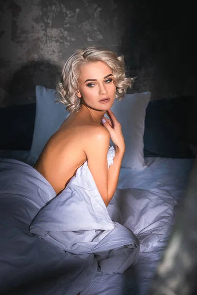 Приваблива гола молода жінка сидить у ліжку під променями заходу сонця, прикриваючи її тіло ковдрою і дивлячись — Stock Photo