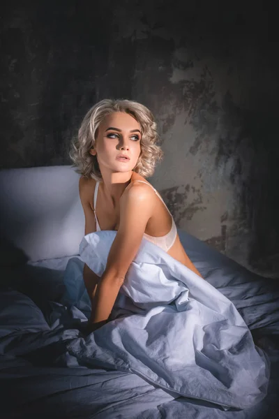 Atractiva joven en ropa interior sentada en la cama bajo los rayos del atardecer y mirando hacia otro lado - foto de stock