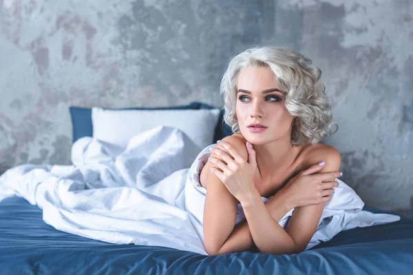 Seductive young woman sitting in bed covered with white blanket — Stock Photo