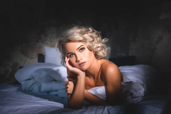 Hermosa mujer joven acostada en la cama cubierta con manta blanca bajo los rayos del atardecer - foto de stock