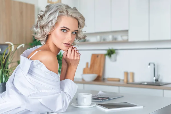 Atractiva joven en camisa blanca con la tableta y la taza de café en la cocina por la mañana - foto de stock