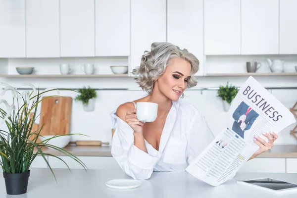 Attraente giovane donna in camicia bianca leggere giornale con caffè in cucina al mattino — Foto stock