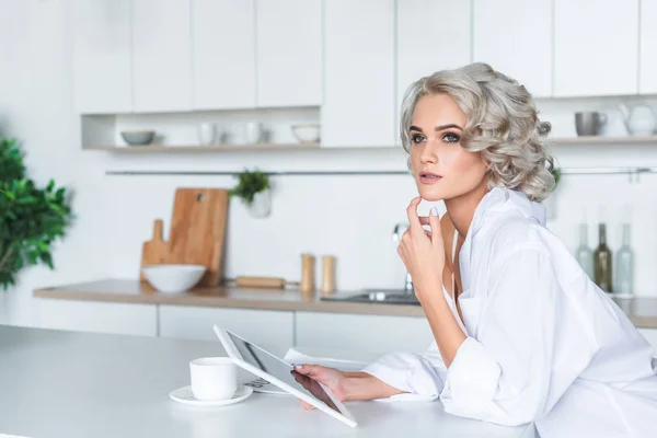 Приваблива молода жінка в білій сорочці тримає планшет і дивиться на кухню вранці — стокове фото