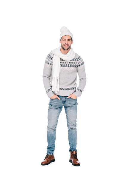 Full length view of handsome young man in sweater, scarf and hat standing with hands in pockets and smiling at camera isolated on white — Stock Photo