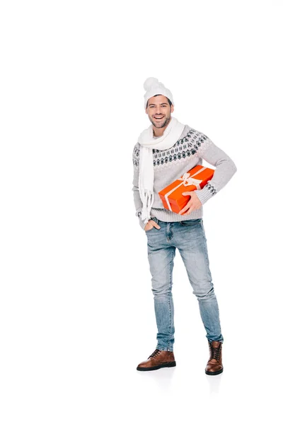 Vista completa de hombre joven guapo en suéter, bufanda y sombrero sosteniendo caja de regalo y sonriendo a la cámara aislada en blanco - foto de stock