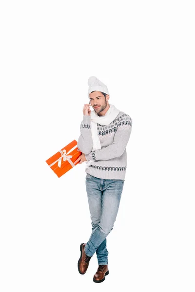 Усміхнений молодий чоловік в светрі, шарфі і капелюсі тримає подарункову коробку і розмовляє по смартфону ізольовано на білому — стокове фото