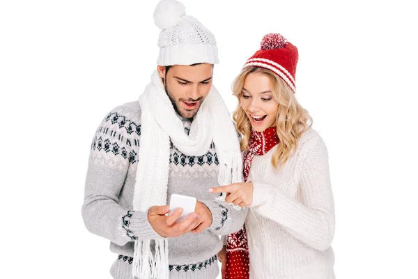 Hermosa pareja joven sonriente en suéteres, bufandas y sombreros usando teléfono inteligente aislado en blanco - foto de stock