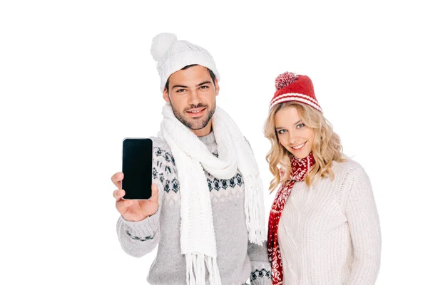 Hermosa pareja joven en traje de invierno celebración de teléfono inteligente con pantalla en blanco y sonriendo a la cámara aislada en blanco - foto de stock