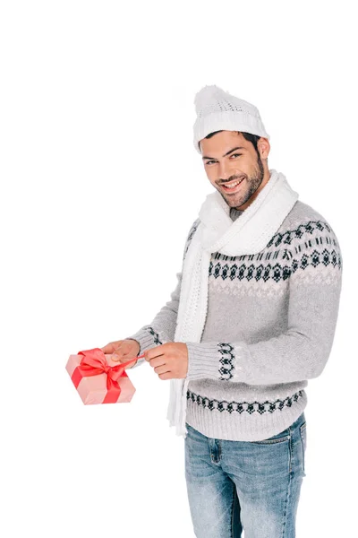 Handsome young man in sweater, scarf and hat opening gift box and smiling at camera isolated on white — Stock Photo