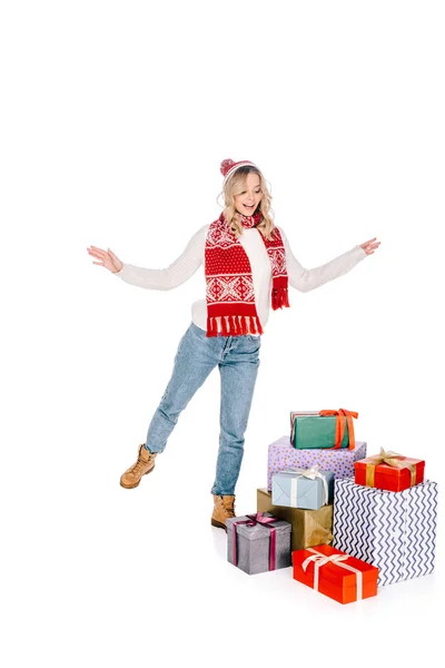 Hermosa mujer joven feliz en bufanda y sombrero mirando cajas de regalo apiladas aisladas en blanco - foto de stock