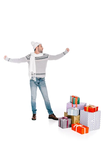 Beau jeune homme en écharpe et chapeau debout près de boîtes-cadeaux empilées isolé sur blanc — Photo de stock