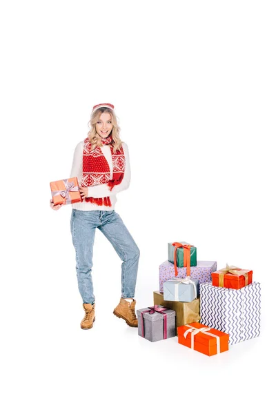 Hermosa mujer joven sosteniendo caja de regalo y sonriendo a la cámara mientras está de pie cerca de regalos apilados aislados en blanco - foto de stock