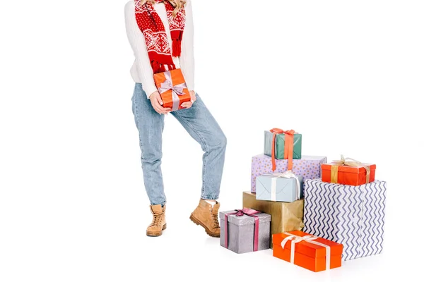 Tiro recortado de mujer joven sosteniendo caja de regalo y de pie cerca de regalos apilados aislados en blanco - foto de stock