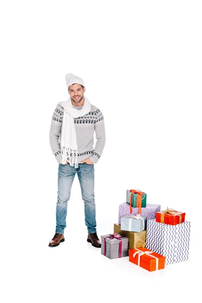 Bello uomo in sciarpa e cappello in piedi con le mani in tasche vicino scatole regalo e sorridente alla fotocamera isolata su bianco — Foto stock